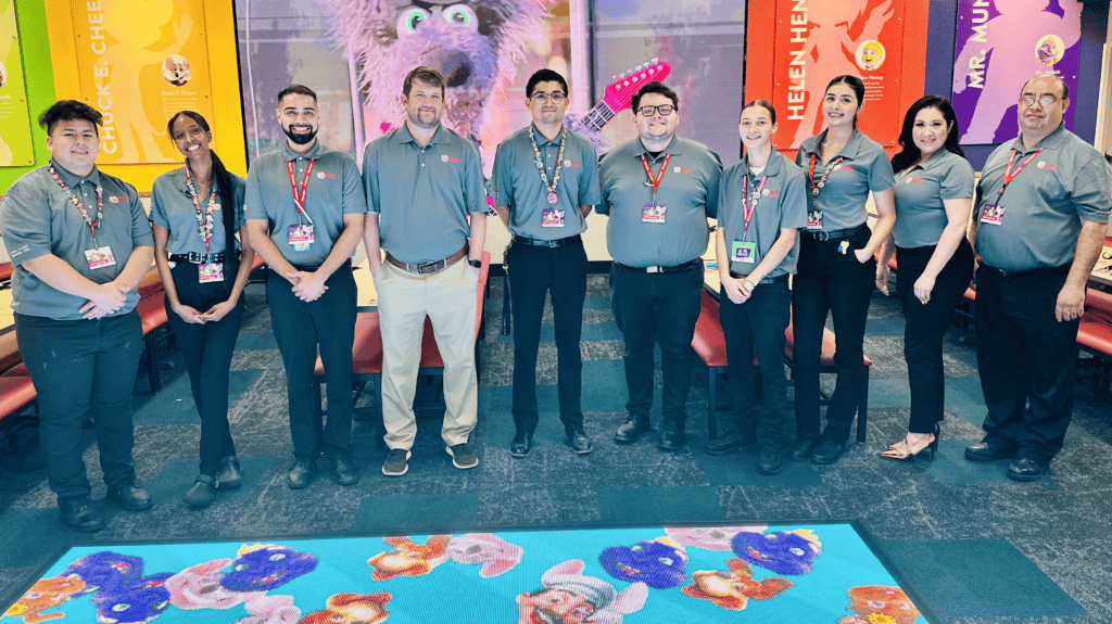 Chuck E. Cheese team, mostly young adults, stand in a party room with character posters and vibrant carpet at the family fun center.