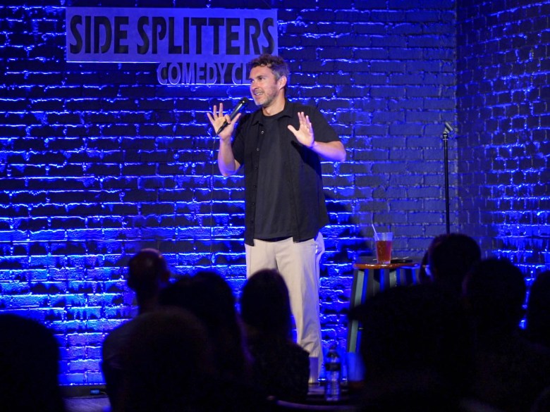 A photograph showing stand-up comedian Mark Normand performing on stage at Side Splitters Comedy Club in Tampa, Florida, in July 2026. Positioned center frame in a full-length shot, Normand gestures outward with both hands while speaking into a microphone. He wears a dark short-sleeve button-down shirt over a dark t-shirt paired with light grey trousers. Behind him, a blue-lit brick wall features a illuminated neon sign reading "SIDE SPLITTERS COMEDY CLUB," with a small stool holding a drink standing to his left and the dark silhouettes of an audience seated in the foreground.