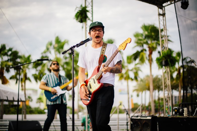 A live outdoor concert photograph by Anthony Martino showing the Tampa indie rock band Brother Cephus performing on stage at the Gasparilla Music Festival in Kiley Garden in Tampa, Florida, on March 11, 2018. Positioned center stage under bright daylight, the lead vocalist Seth Davis and guitarist sings passionately into a microphone while playing a red electric guitar with a white pickguard. They wear a plain white short-sleeve T-shirt, dark pants, round glasses, and a green baseball cap, with visible arm tattoos. In the midground to the left, a second guitarist, Gabe Davis, wearing sunglasses and a blue-and-white vertically striped short-sleeve shirt plays a white electric guitar, framed by tall palm trees and stage trusses against a soft cloudy sky.