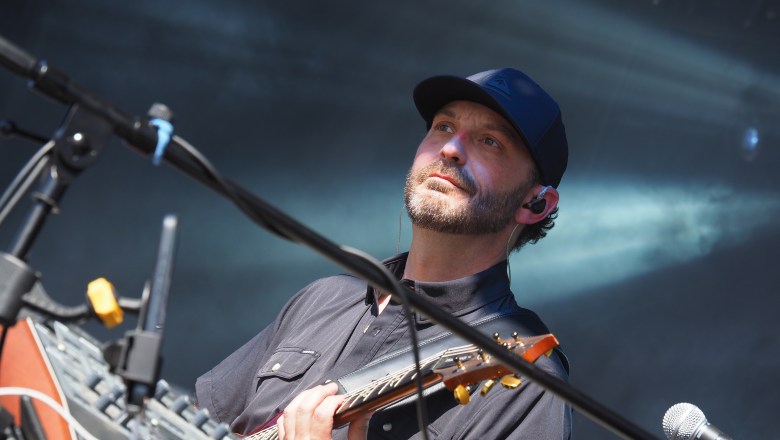 A live music photograph of a musician from the band MiniM performing on stage at the Gasparilla Music Festival at Meridian Fields in Tampa, Florida, on April 22, 2026. The artist is framed from the chest up, looking upward and off-camera with a calm expression. They have a short beard and are wearing a dark blue baseball cap, a dark short-sleeve button-down shirt, and an in-ear monitor. The musician is holding an electric stringed instrument with a natural wood finish and gold hardware across their chest. Several black microphone stands cross the foreground, while the background features bright, hazy white and blue stage lighting.