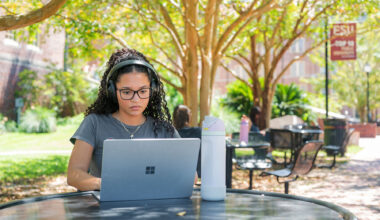 A young woman with glasses focuses on her laptop while sitting outside on campus.