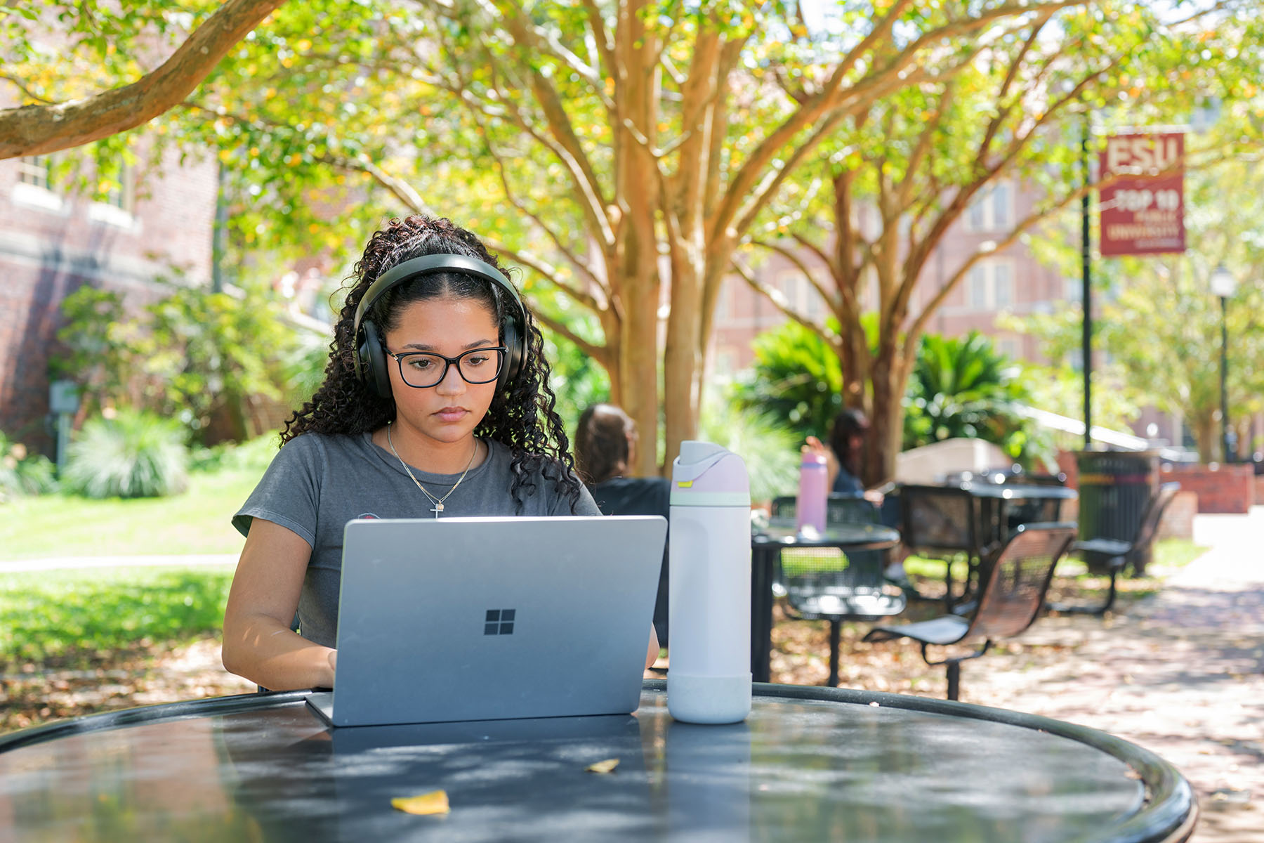 A young woman with glasses focuses on her laptop while sitting outside on campus.