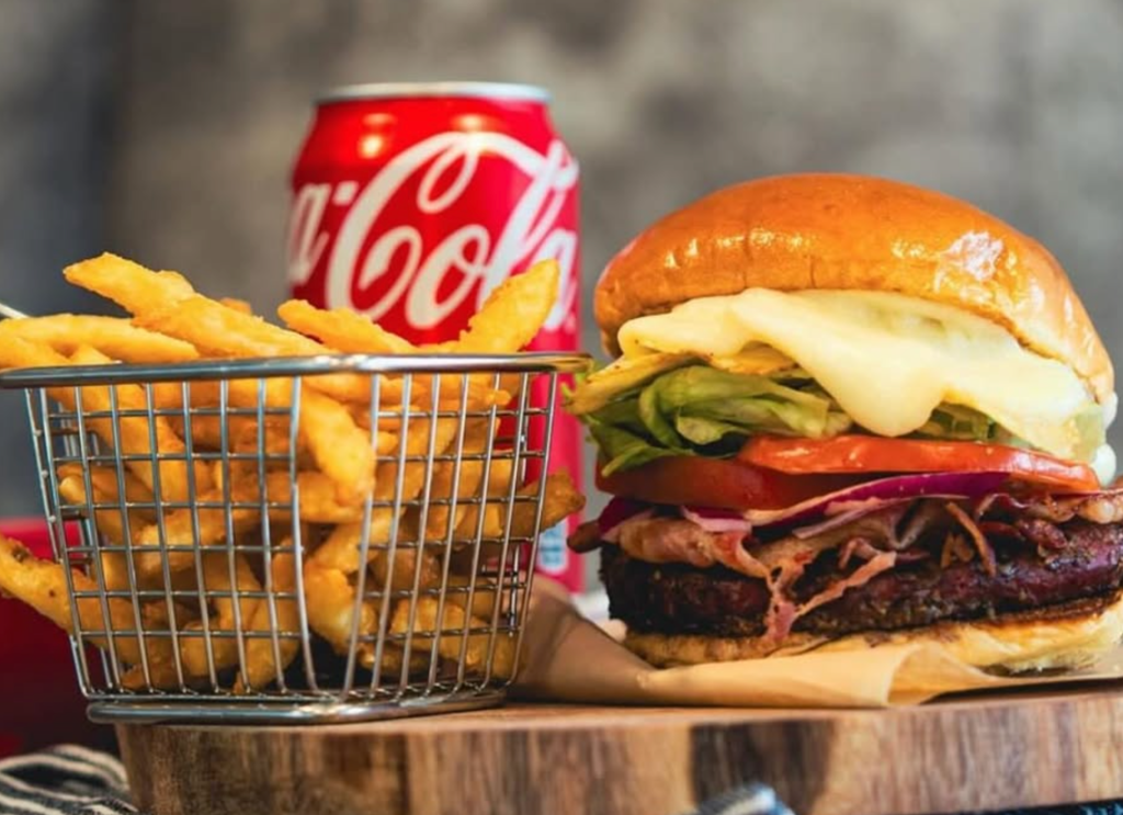a loaded cheeseburger with toppings next to a tray of French fries and a Coca-Cola can