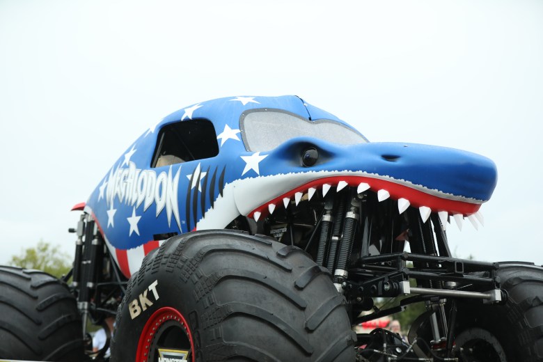 A low-angle close-up photograph showing the Megalodon Stars and Stripes monster truck on display at a Walmart in Tampa, Florida, on July 17, 2026. Custom-designed to resemble a 3D shark with a red, white, and blue patriotic paint scheme, the truck features large white stars across its blue fiberglass body, 3D shark teeth lining its gaping open jaw, and the "MEGALODON" logo in stylized silver lettering on its side panel. Massive black BKT tires with red rim accents and heavy-duty suspension shocks frame the lower portion of the vehicle against a bright outdoor sky background.