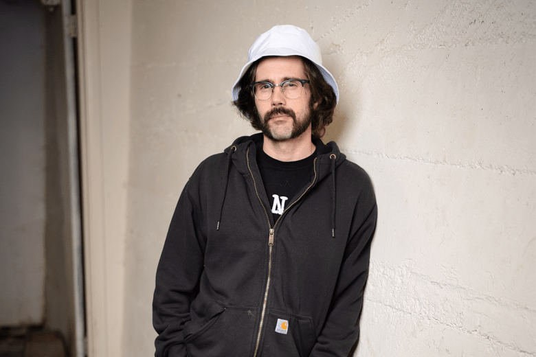 A press portrait photograph of stand-up comedian Nick Mullen leaning against a light-colored wall indoors. Captured in a medium three-quarter shot, Mullen stands centered while wearing a light grey bucket hat, clear wire-rimmed glasses, a dark zip-up hoodie over a dark t-shirt, and dark pants. He features dark shoulder-length hair and a full mustache, standing in a relaxed pose against a minimalist interior backdrop with soft overhead lighting.
