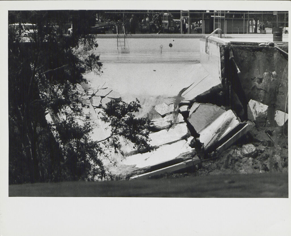 Black & white photo of swimming pool with one side collapsed into sinkhole
