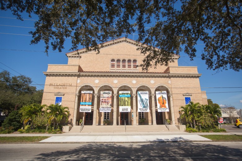 An exterior photograph of The Palladium Theater in St. Petersburg, Florida. Captured from across the street at a low angle, the historic 1925 Mediterranean and Romanesque Revival-style building features a beige brick facade, a grand grand entrance with three arched doorways set under white stone archways, and decorative wrought-iron light fixtures mounted between the arches. Above the entrance, a tiled decorative band and small arched windows accent the second story, topped by a red-barrel tile roof line. Black metal railings frame the front steps leading to the main entrance along a clean concrete sidewalk
