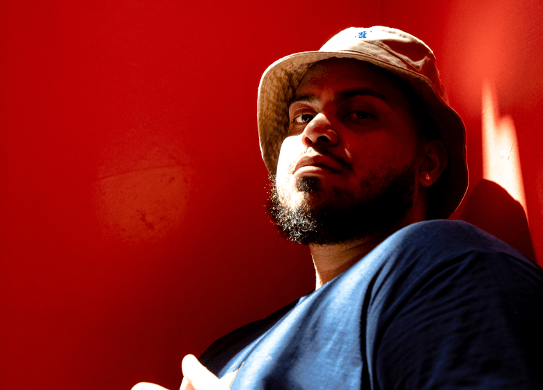 A low-angle portrait photograph by Roberto Rivera showing Tampa rapper Perception posing against a bold red background under dramatic lighting. Positioned center frame in a medium close-up, Perception looks down toward the camera with a confident expression and a full dark beard. He wears a light tan bucket hat angled slightly over his forehead and a blue t-shirt, highlighted by a strong warm light source casting high-contrast shadows across the left side of his face.