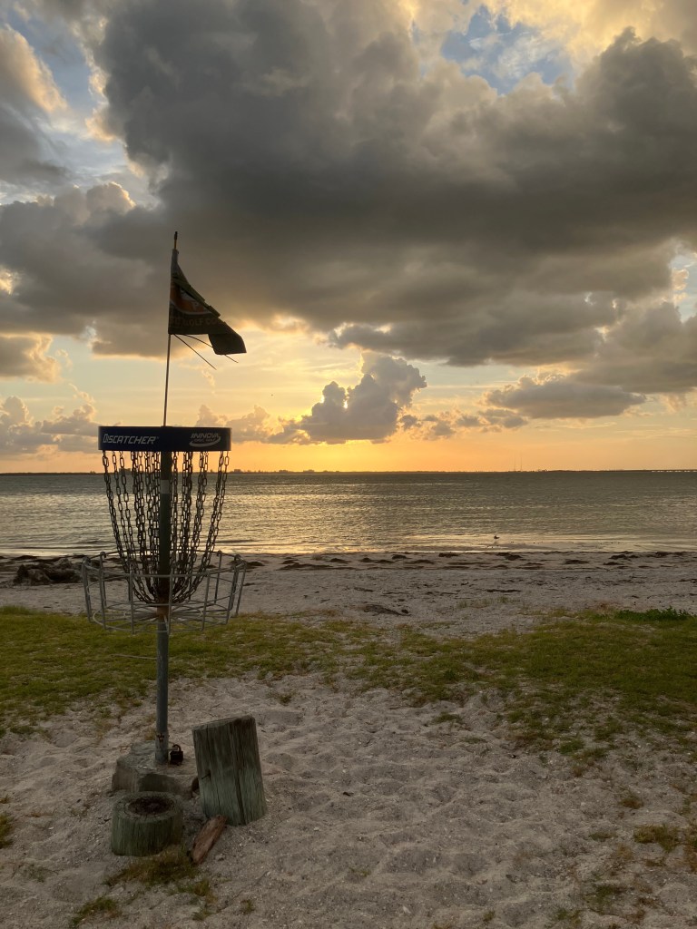 A scenic sunset photograph of a metal disc golf basket standing on the sandy beach at Picnic Island Park in Tampa, Florida, on June 25, 2026. The basket, which features a small dark flag blowing in the wind on top, is positioned in the foreground amidst patches of grass and sand, with small wooden posts at its base. In the background, the calm waters of the bay stretch toward a distant horizon under a dramatic sky filled with thick, heavy gray clouds. A warm, golden light from the setting sun breaks through the clouds near the horizon, reflecting softly on the water.