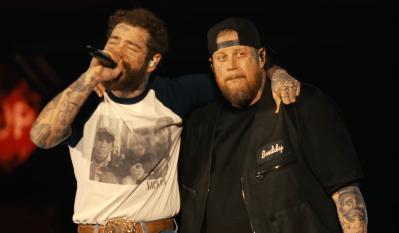 A live music screenshot of musicians Post Malone and Jelly Roll standing closely together on stage during a performance at T-Mobile Park in Seattle, Washington on June 26, 2025. On the left, Post Malone, who has extensive face and arm tattoos, sings into a microphone while wearing a white raglan shirt with dark sleeves featuring a band graphic, paired with a large gold belt buckle. Post Malone has one arm draped affectionately over the shoulder of Jelly Roll on the right. Jelly Roll, also sporting face tattoos and a beard, looks forward wearing a backwards black baseball cap, a black t-shirt, and a black overshirt with the word "Buddy" embroidered on the chest.