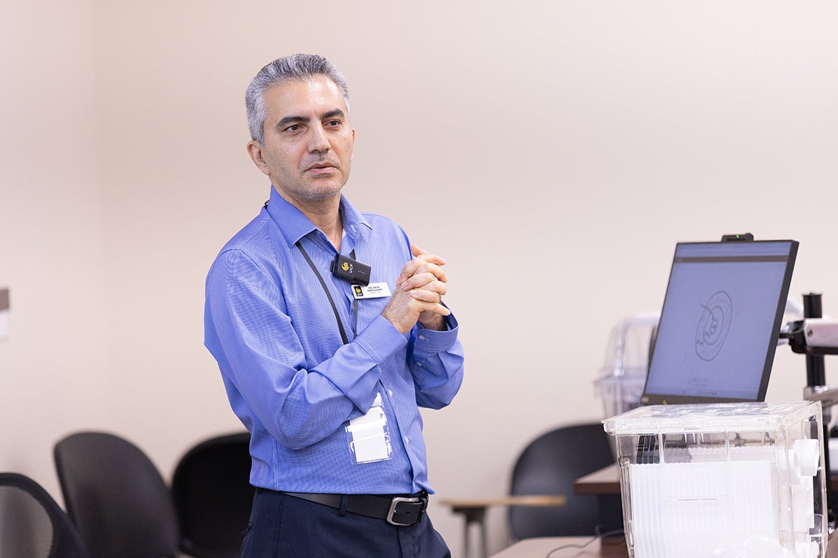 Gray-hair man in blue long sleeve collar shirt stands with hands clasped in front of him next to a screen