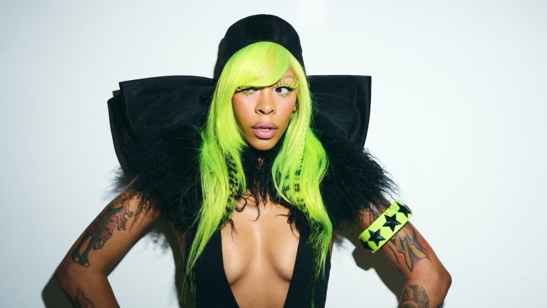 A medium shot of artist Rico Nasty with vibrant, neon green hair and a black cap, wearing a structural black outfit with a dramatic structured collar. She poses against a plain white background, looking off-camera.