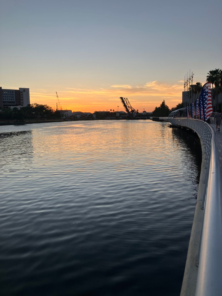 A scenic twilight photograph looking down the Tampa Riverwalk in Tampa, Florida, taken on June 23, 2026. A curved, paved pedestrian walkway with a smooth metal railing sweeps along the right side of the frame, bordering a wide, calm river. The path is lined with palm trees and several tall American flag banners. In the distance, the dark silhouette of the Cass Street Bridge spans the water, set against a clear sky glowing with warm orange, peach, and soft blue sunset hues that reflect gently on the water's surface. A single individual can be seen walking down the path away from the camera.