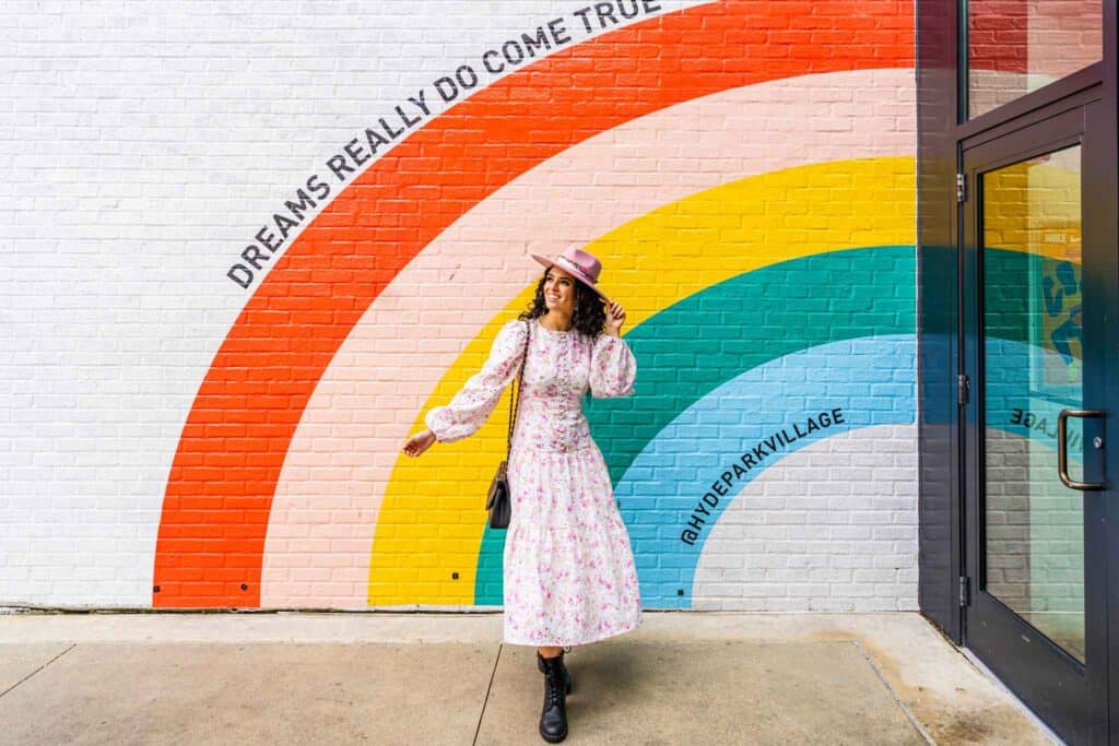 woman in a dress and hat poses in front of a colorful rainbow wall
