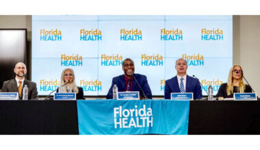 Five panelists, three men and two women, are seated at a table with microphones and water bottles, in front of a backdrop and table banner displaying the 'Florida HEALTH' logo.