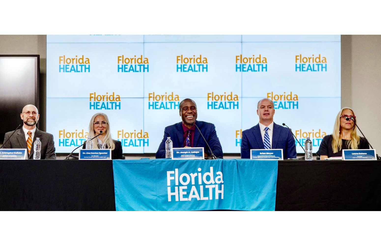 Five panelists, three men and two women, are seated at a table with microphones and water bottles, in front of a backdrop and table banner displaying the 'Florida HEALTH' logo.