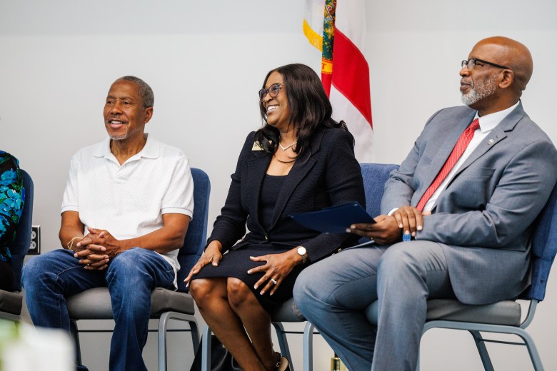 A photograph of State Senator Darryl Rouson, Dr. Tonjua Williams, and Mayor Ken Welch seated together at the SPC Gibbs Campus in St. Petersburg, Florida, on September 29, 2023. On the left, Sen. Darryl Rouson sits with hands clasped, wearing a short-sleeve white polo shirt and blue jeans. In the center, Dr. Tonjua Williams smiles broadly, wearing a dark suit, glasses, and a necklace, while holding a dark folder. On the right, Mayor Ken Welch looks off-camera to the left, wearing a grey suit jacket, a white button-down shirt, a bright red tie, and glasses, with documents resting on their lap. The three individuals are seated on blue padded chairs against a plain white wall, with a Florida state flag partially visible in the background.