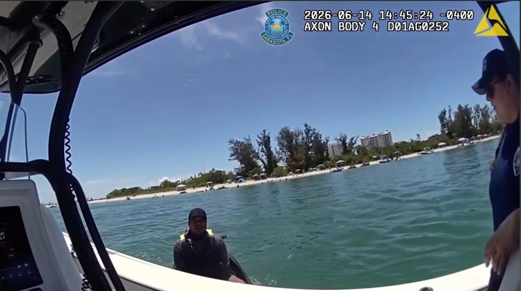 Bodycam footage from a police boat on a sunny day shows an officer and a man in a life vest on a jet ski, with a beach and buildings in the background.