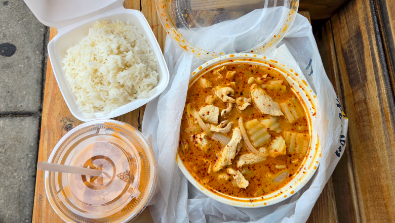 A top-down overhead photograph of a takeout Thai meal from Chalawan Thai Delish in Tampa, Florida, on July 16, 2026, set on an outdoor wooden bench next to a concrete sidewalk. In the center, a round clear plastic bowl filled with rich orange Massaman curry sits lined in a white plastic bag, featuring visible pieces of sliced chicken, potatoes, and onions topped with red chili flakes. To the upper left, an open small square styrofoam container holds steamed white Jasmine rice. To the lower left rests a plastic cup filled with iced orange Thai iced tea, complete with a straw inserted through the lid.