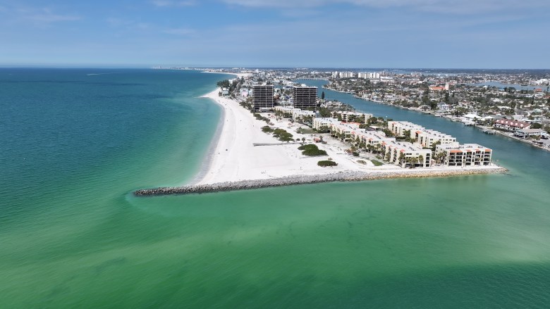An aerial drone photograph overlooking the coastline of St. Pete Beach, Florida, on a bright, sunny day. In the foreground, vibrant emerald green water meets a stretch of bright white sand that extends outward, bordered by a long, rocky breakwater jetty. Along the shoreline, mid-rise condominiums and resort buildings line the beach, while a calm intracoastal waterway runs parallel on the right side of the frame, lined with coastal homes and boat docks. The scene stretches toward the horizon under a clear, expansive blue sky.