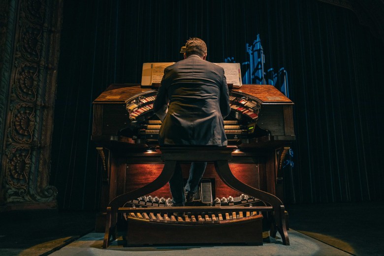 A photograph by Javi Ortiz showing organist Dr. Steven Ball performing on the Mighty Wurlitzer Theatre Organ at the historic Tampa Theatre in Tampa, Florida, on March 13, 2021. Captured from behind in a wide-angle shot, Dr. Ball sits centered on the wooden organ bench facing the elaborate, curved horseshoe console of the organ, which features glowing red accent lights and multiple horseshoe ranks of colorful stop tabs and keys. Dr. Ball wears a dark grey suit jacket and glasses while playing, set on a dark stage against a dramatic black curtain backdrop flanked by ornate architectural columns.