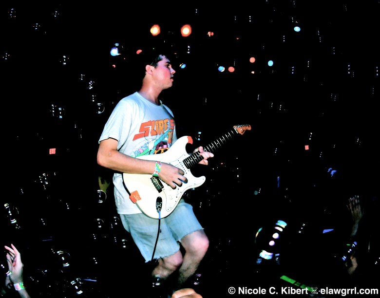 A live concert photograph by Nicole C. Kibert showing John Paul Pitts of Surfer Blood performing on stage at the State Theatre in St. Petersburg, Florida, on September 19, 2010. Captured in profile facing right against a dark stage background, the lead guitarist and vocalist plays a white Fender Stratocaster electric guitar while standing on stage. They are wearing a light-colored graphic T-shirt with a colorful vintage print and light denim shorts. Floating soap bubbles catch the stage lights around them against the dark background, with stage lights glowing softly overhead and the hands of audience members visible in the lower foreground.