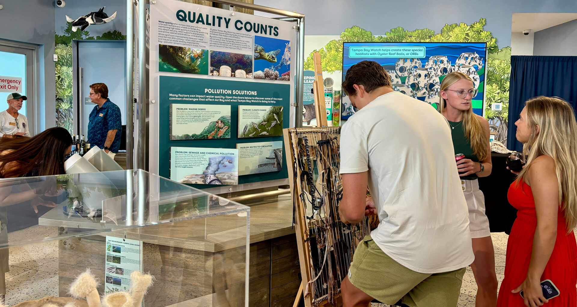 Three young adults interact with artwork and an indoor educational exhibit focused on environmental quality and pollution.