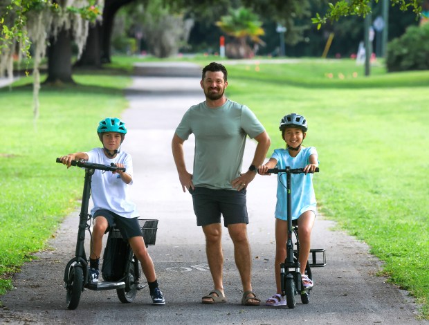 Siblings Max Buza,11, and Casey Clark, 10, with dad Nate Clark with their electric scooters on Cady Way Trail in in Orlando, Friday, July 24, 2026. During the school year, the students use their e-scooters to commute 1.4 miles daily to Baldwin Park Elementary School. The Orange County School Board will vote Tuesday on a proposal to ban e-bikes and e-scooters at all elementary and middle school campuses for students without a driver's license beginning in the 2026-27 school year. (Joe Burbank/Orlando Sentinel)