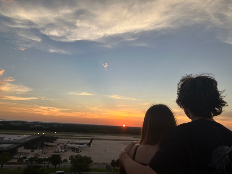 A scenic sunset photograph overlooking the tarmac at Tampa Airport in Tampa, Florida, taken on June 28, 2026. In the foreground, the dark silhouettes of two individuals stand close together, viewed from behind, with one person resting an arm around the other's back as they look out toward the horizon. Below them, the expansive airport grounds feature parked passenger airplanes, including one with a visible Delta tail fin, situated near terminal buildings and ground equipment. In the background, a bright orange sun sets on the distant horizon under a vast sky filled with textured clouds reflecting soft twilight hues.
