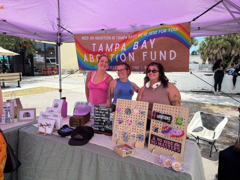 A photograph of three volunteers standing behind a Tampa Bay Abortion Fund outreach table under a purple canopy tent outdoors in St. Petersburg, Florida. Hanging at the back of the tent is a colorful banner with a rainbow border that reads "NEED AN ABORTION IN TAMPA BAY? WE'RE HERE FOR YOU! TAMPA BAY ABORTION FUND." The three individuals stand side-by-side smiling toward the camera; from left to right, one wears a bright pink tank top, the center person wears a blue tank top and glasses, and the individual on the right wears a mauve top, sunglasses, and white headphones around their neck. The table in front of them is draped in a grey tablecloth and covered with advocacy materials, including hats, a small chalkboard sign, stickers displayed on wooden pegboards, and literature. The background shows an open outdoor plaza with buildings, palm trees, and a few bystanders under bright daylight.
