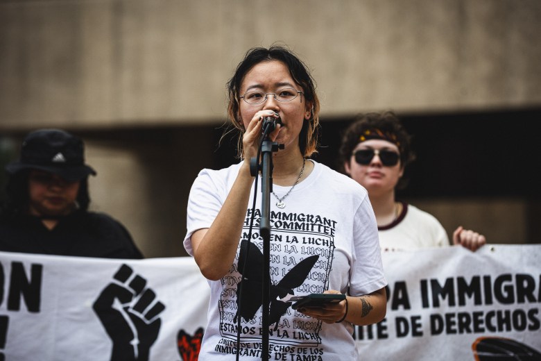 A photograph of a member of the Tampa Immigrants Rights Committee speaking at a microphone during an ICE protest outside Tampa City Hall in Tampa, Florida, on July 19, 2026. Positioned center stage, the speaker is framed from the waist up, wearing clear glasses and a white t-shirt featuring a large black butterfly design and printed text that reads "TAMPA IMMIGRANT RIGHTS COMMITTEE / UNIDOS EN LA LUCHA." They are holding a microphone in one hand while speaking passionately. Behind them, two other individuals stand supporting a white banner; the person on the left wears a dark bucket hat and black shirt, while the person on the right wears sunglasses. The banner features a black raised fist icon and text, with part of the phrase "A IMMIGRANT RIGHTS COMMITTEE" clearly visible. The background consists of a brutalist, light grey concrete building facade under soft daylight.