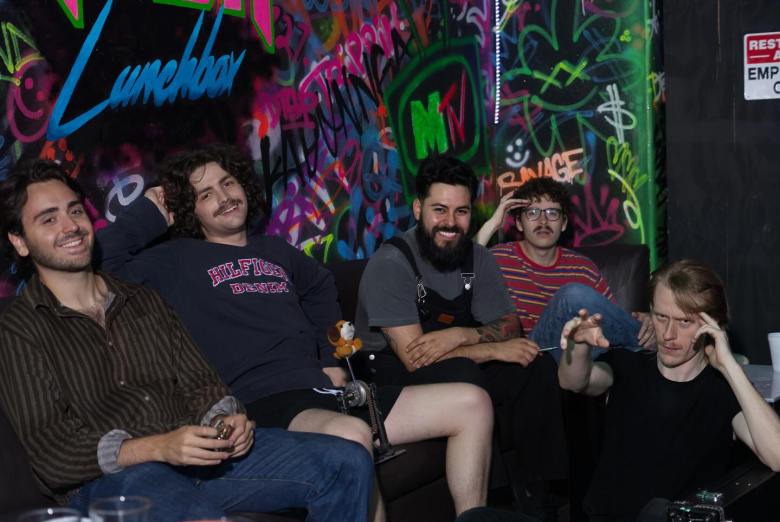 A photograph by Adam Boehm showing the five members of Tampa band The George sitting together on a dark couch inside a brightly decorated venue space. From left to right, the band members pose in casual attire—including a striped button-down shirt, long-sleeve graphic tops, overalls, and a red-and-blue striped shirt—smiling and leaning comfortably against one another. Behind them, a colorful graffiti wall displays vibrant artwork, neon-style signage reading "Lunchbox," and a bootleg lookin ass "MTV" logo, set under dim interior lighting.