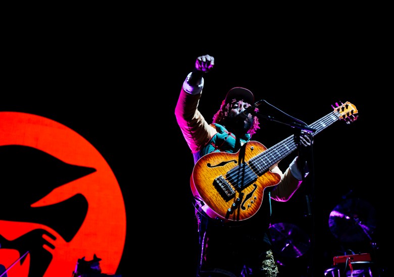 A live music photograph by Maria Jose Govea of musician Thundercat performing on stage at the Red Bull Music Academy Festival in Los Angeles, California, on Oct. 14, 2017. The artist is framed from the waist up, passionately playing an ornate, sunburst-finished six-string semi-hollow body bass guitar. They have dark braided hair and are wearing a maroon jacket featuring green and yellow shoulder panels. The dramatic stage lighting casts a magenta glow on the performer, who is positioned in front of a striking background featuring a large, glowing red circular graphic with a black silhouette design set against the dark stage.
