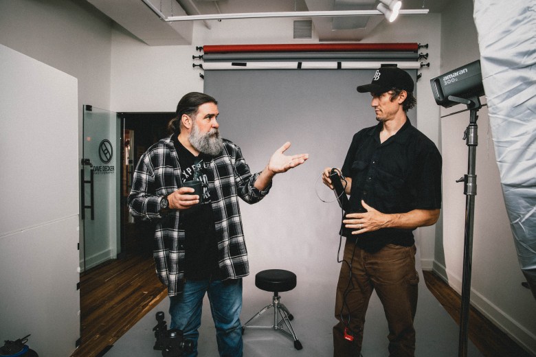 A wide-angle interior photograph showing Crowbar owner Tom DeGeorge and filmmaker Tyler Riggs standing inside the Dave Decker Photography Studio in Ybor City, Florida, on March 25, 2026. On the left, Tom DeGeorge stands with long hair tied back and a full grey beard, wearing an unbuttoned black-and-white plaid flannel shirt over a dark graphic t-shirt, blue jeans, and dark boots while holding a camera and gesturing outward with his left hand. On the right, Tyler Riggs stands facing him with a black baseball cap, a dark short-sleeve button-down shirt, brown trousers, and dark shoes, holding camera gear with both hands. Set in a photography studio, the background features studio light stands, softboxes, a grey paper backdrop roll with red and blue paper accents, and a swivel stool.
