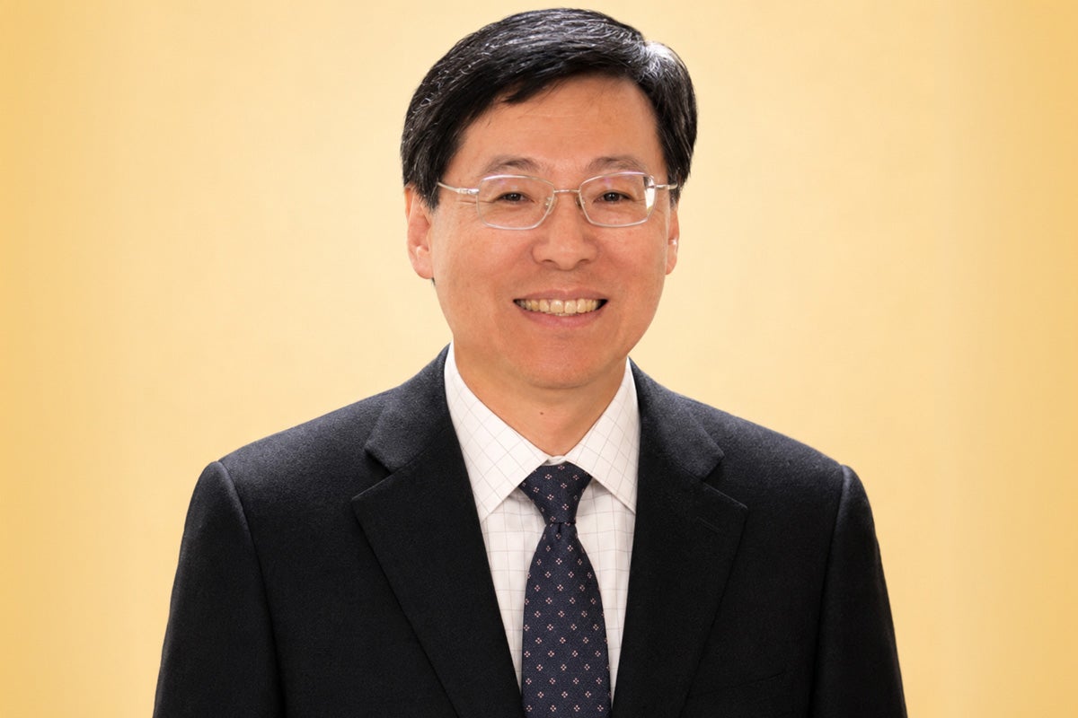 Portrait of smiling Asian man wearing glasses and black business jacket with white shirt and black tie in front of yellow backdrop