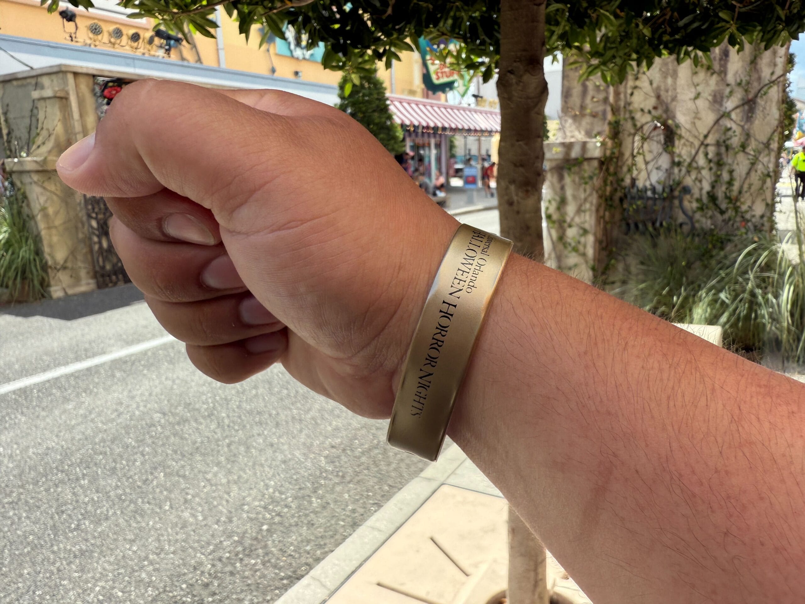 A hand wearing a gold Wristband System with engraved text, held up outdoors near shops during Halloween Horror Nights 2025.