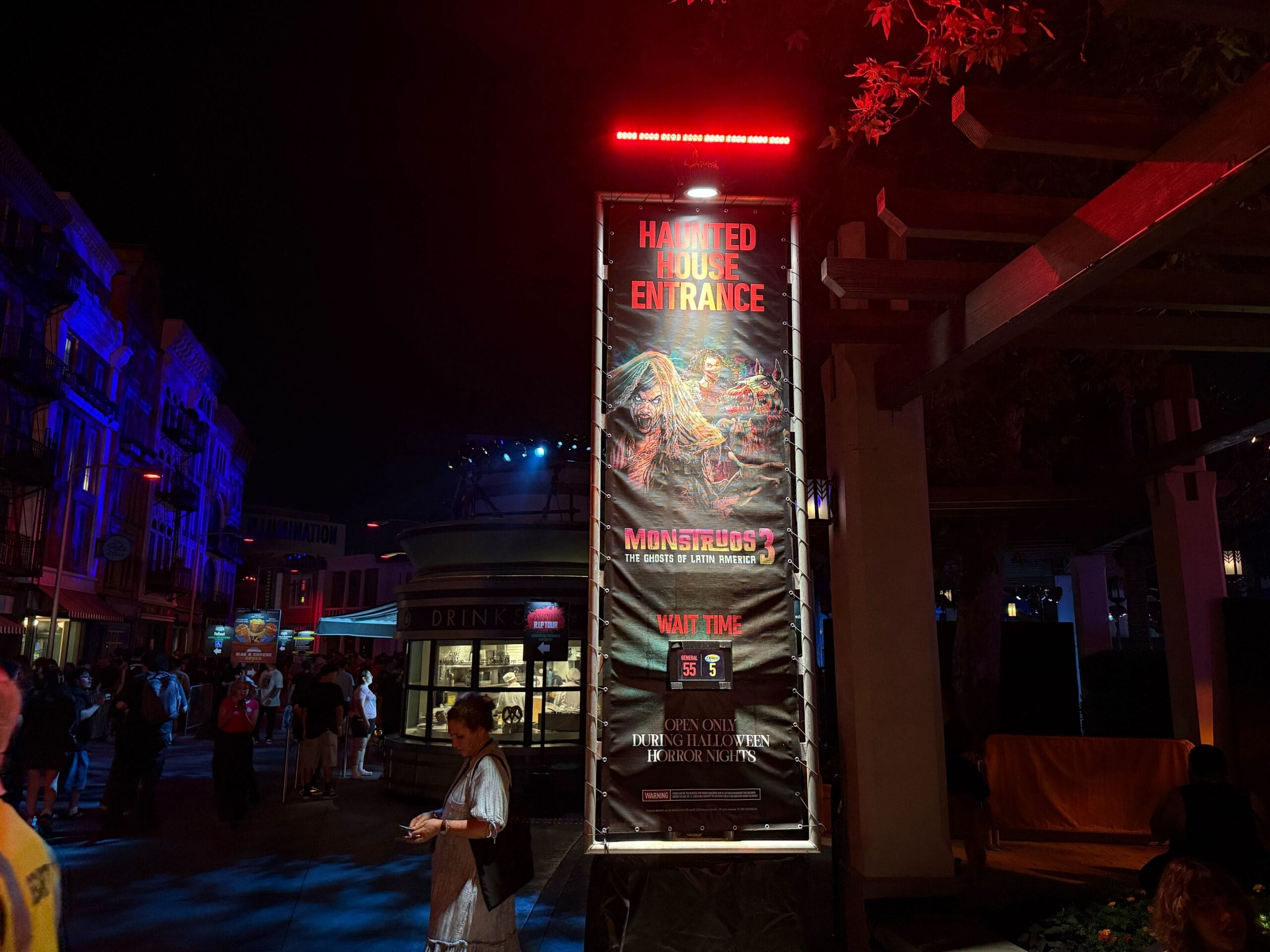 A haunted house entrance sign at night for USH HHN 2025, showing Monstros 3 wait time and a monster graphic nearby.