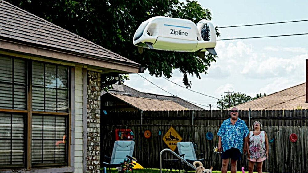 A Zipline autonomous drone lowers a medical delivery into a residential backyard as two homeowners watch from beside their swimming pool.