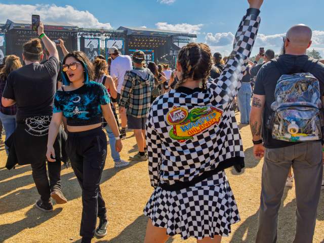 Festival attendees gather in front of a main stage during Vans Warped Tour Orlando at Tinker Field on the Camping World Stadium Campus. A person in the foreground wearing checkered Vans apparel raises an arm toward the stage while others record and watch the live performance.