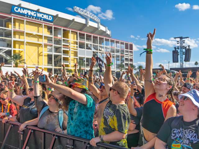 A dense crowd of festival attendees raises hands toward a stage during Vans Warped Tour Orlando at the Camping World Stadium Campus. The stadium structure and event production elements are visible in the background.