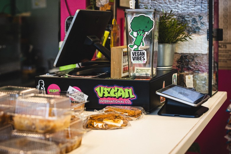 An interior photograph of the checkout counter at Vegan International Co. Market & Cafe in Tampa, Florida. Taken from a close-up angle, the counter displays a black cash register terminal alongside a clear tip jar decorated with a cartoon broccoli sticker reading "VEGAN VIBES." Below the register, the counter front features a colorful sticker with stylized purple-and-yellow lettering that reads "VEGAN INTERNATIONAL CO." In the foreground, packaged baked goods and cookies in clear plastic containers rest on the counter next to a digital payment tablet.