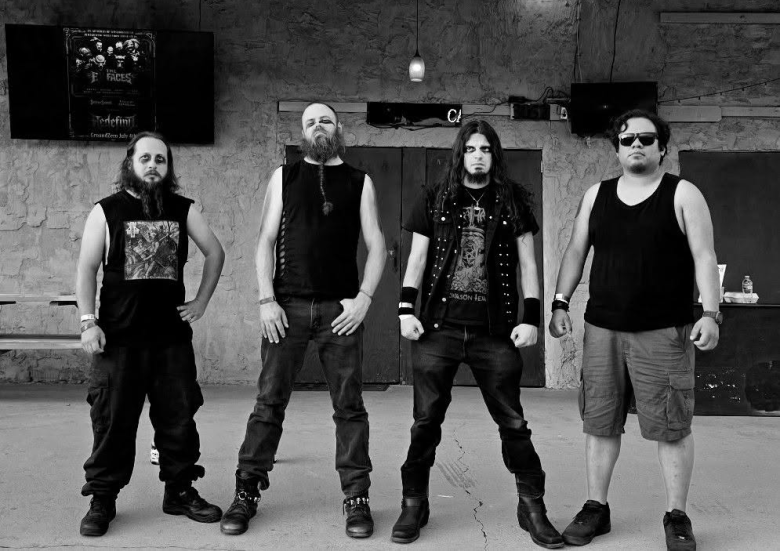 A black-and-white portrait photograph of the three members of Tampa black metal band Voidrium standing outdoors in front of the Ground Zero music venue. The three band members stand side-by-side wearing black metal corpse paint around their eyes. On the left, a member stands with hands on hips wearing a dark sleeveless graphic shirt. In the center, a member with a long braided beard stands with hands in his pockets wearing a sleeveless shirt. On the right, a member with long dark hair wears a studded denim vest over a graphic shirt. In the background, a hanging string light fixture and concert poster decorate the building wall under a large overhead sign reading "GROUND ZERO."