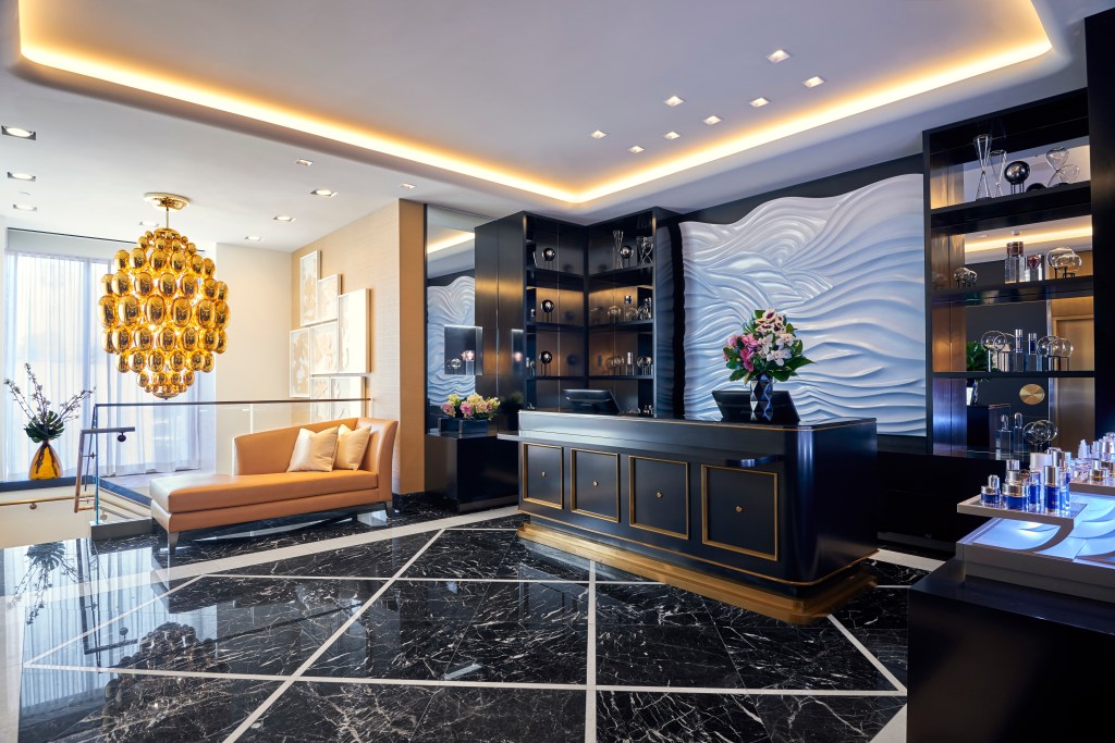 Luxurious reception area at the Waldorf Astoria Beverly Hills with a black marble floor, modern black desk, textured white wall, and a golden chandelier.