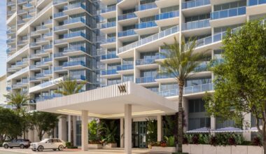 Modern high-rise building with glass balconies and a covered entrance to the Waldorf Astoria Miami Beach surrounded by palm trees and greenery.