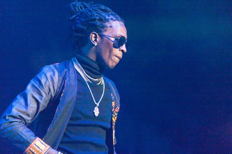 A live music photograph of hip-hop artist Young Thug performing at Philips Arena in Atlanta, Georgia, on December 10, 2016. Framed from the chest up and turned in profile looking downward to the right, the musician has their hair styled back and is wearing dark sunglasses. They are dressed in a black turtleneck under a dark jacket with patterned sleeve details, heavily accessorized with layered silver chains and a prominent hand-shaped pendant. The background consists of a solid, vibrant wash of deep blue stage lighting.