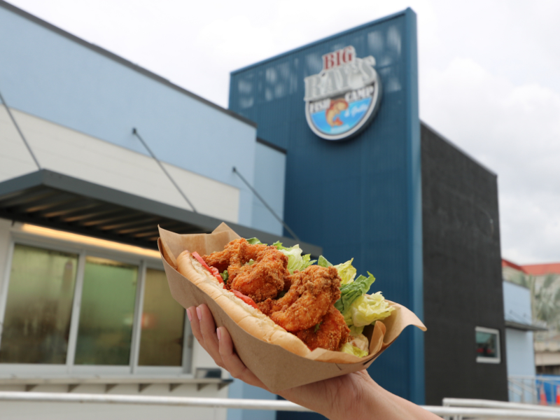 Hand holds up a breaded shrimp po boy with lettuce and tomato on a bun in a cardboard container with the Big Ray's Fish Camp sign in the background.