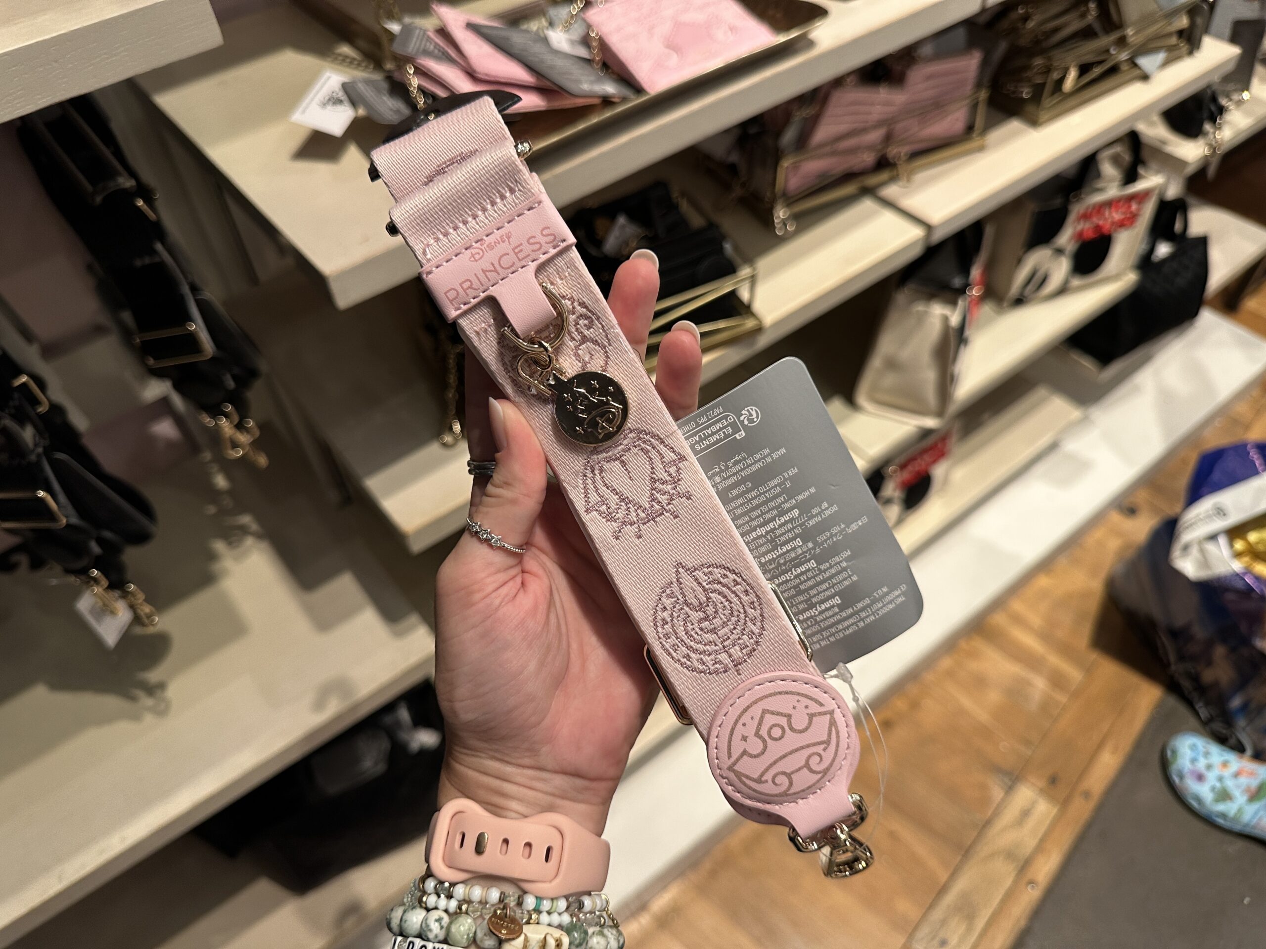 This pink bag strap is Disney Princes themed. It has various princess iconography patterned throughout. A silver-toned charm hangs on it with the Disney