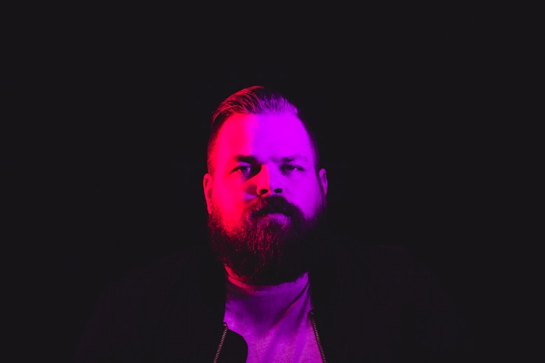 A dramatic, dark promotional portrait by Effixx of electronic music producer Com Truise set against a solid black background. Positioned center frame in a chest-up portrait, the individual looks forward toward the camera with a full dark beard and neatly styled dark hair. Neon lighting illuminates the portrait with a strong dual-color split effect: bright vibrant magenta-pink light casts across the left side of their face and neck, while subtle blue-purple light highlights the right side, with the rest of their dark outfit blending seamlessly into the shadows.