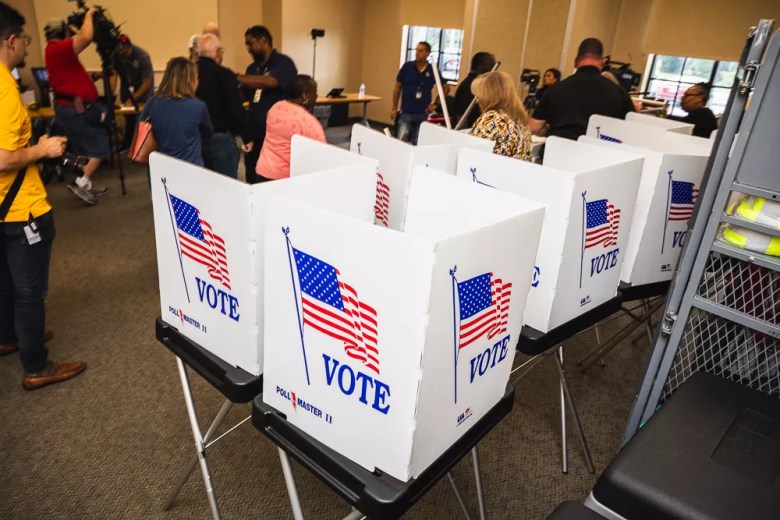 A row of white voting booths with 'VOTE' and American flag graphics inside a busy polling place or election office, with people and camera crew visible in the background.