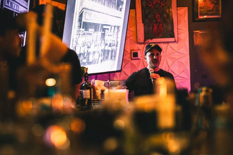 A photograph of speaker Manny Leto giving a presentation at the venue Shuffle in Tampa, Florida, on July 15, 2025. The image is shot with a shallow depth of field, featuring a heavily blurred foreground filled with the silhouettes of audience members, glass bottles, and bar items illuminated by warm bokeh lights. In the sharp midground, the individual stands holding a microphone, wearing a dark short-sleeved t-shirt and a dark baseball cap. Behind them, a bright projector screen displays a black-and-white historical photograph of a storefront and a group of people. The background is dimly lit with pinkish hues, highlighting a wall with a white geometric textured pattern and a framed piece of artwork hanging to the right.